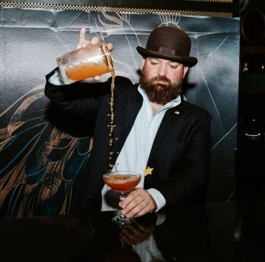 Alumnus Alex Tilkens pouring a cocktail into a glass at his brick and mortar location in Amesbury.