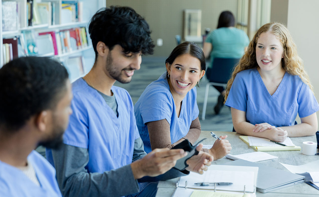 Young, diverse male and female students wearing their blue medical scrubs sitting at a table in the library work together to do their classwork