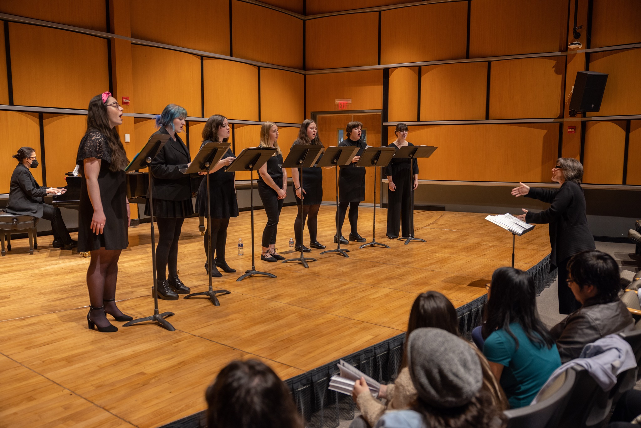 People standing on stage singing dressed in black clothing with music stands in front of them.