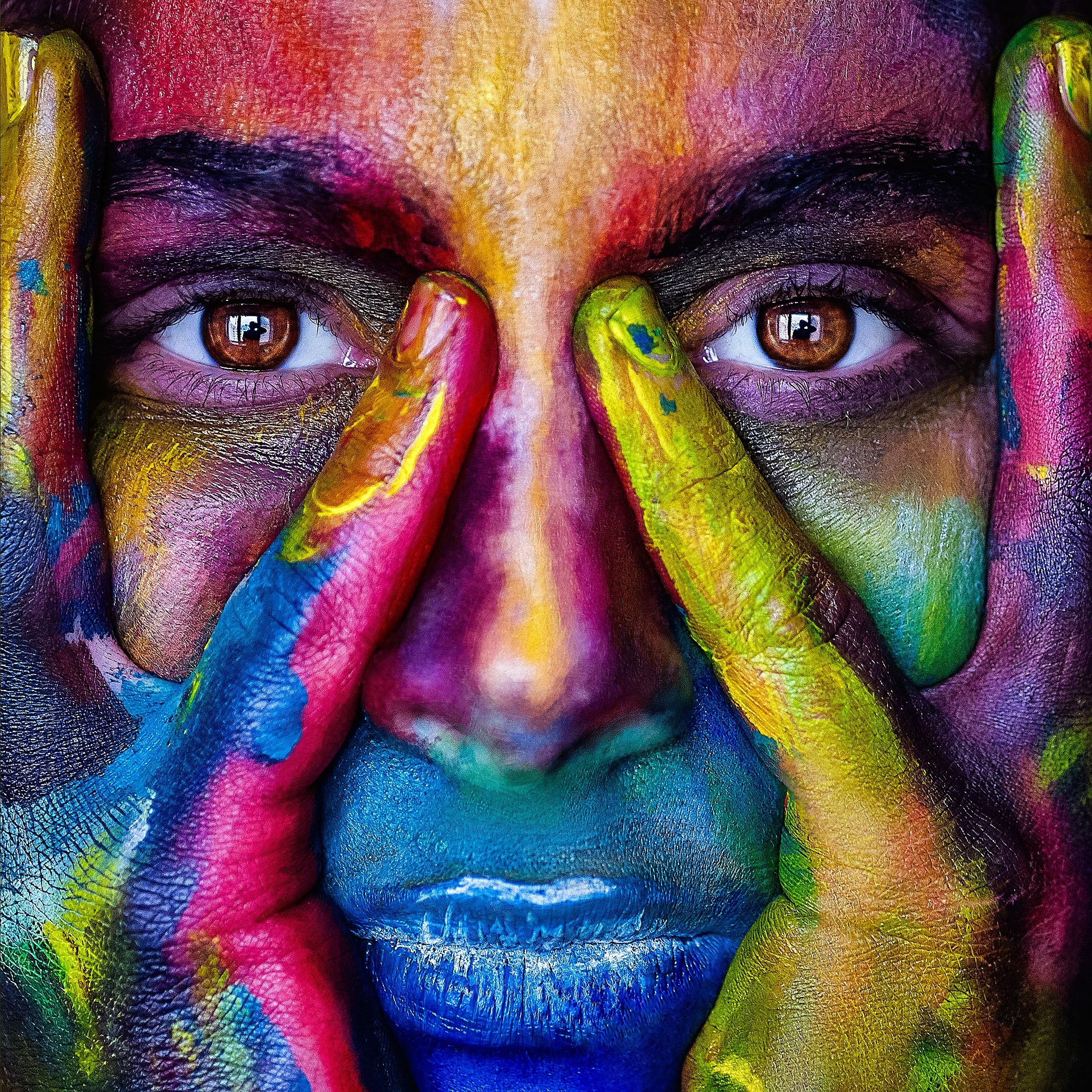 Image of a person's face, covered in paint of all colors. The person is holding their face in their hands and looking directly at the camera.