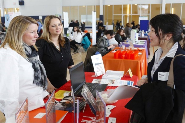 A student in a blazer stands at a table and speaks with two women who are behind the table. They are in an auditorium with other employers at tables.