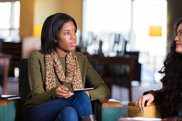 A student in jeans and a sweater sitting in a chair talking to another student