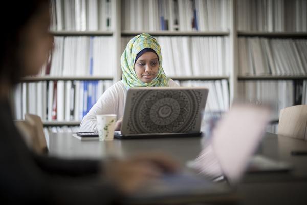 girl wearing headscarf, sitting at a large desk in front of a wall of bookshelves, using her laptop
